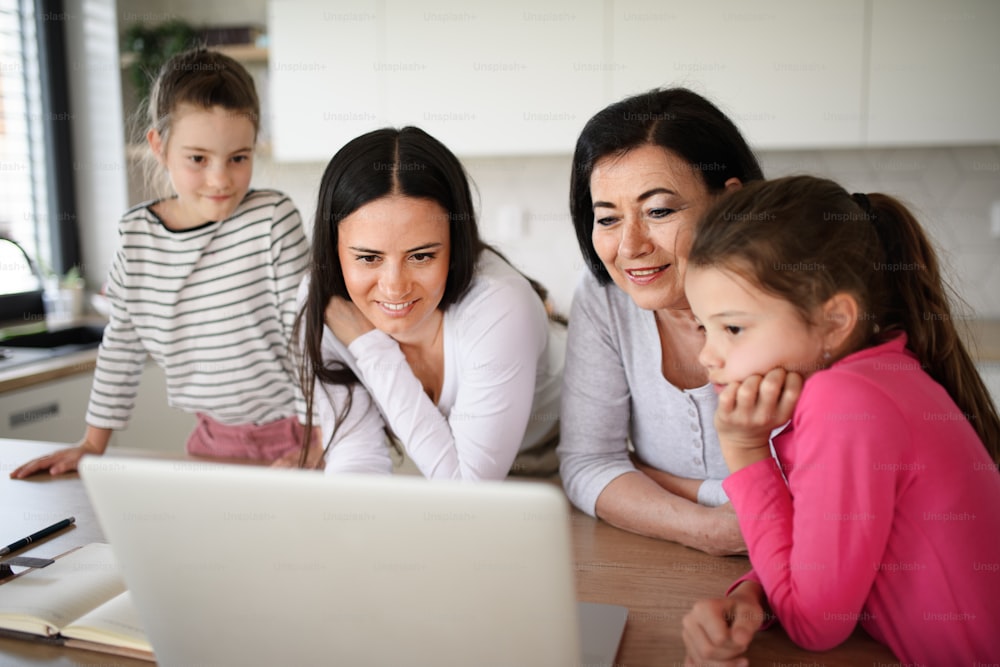 Retrato de niñas pequeñas felices con la madre y la abuela en el interior de la casa, usando una computadora portátil.