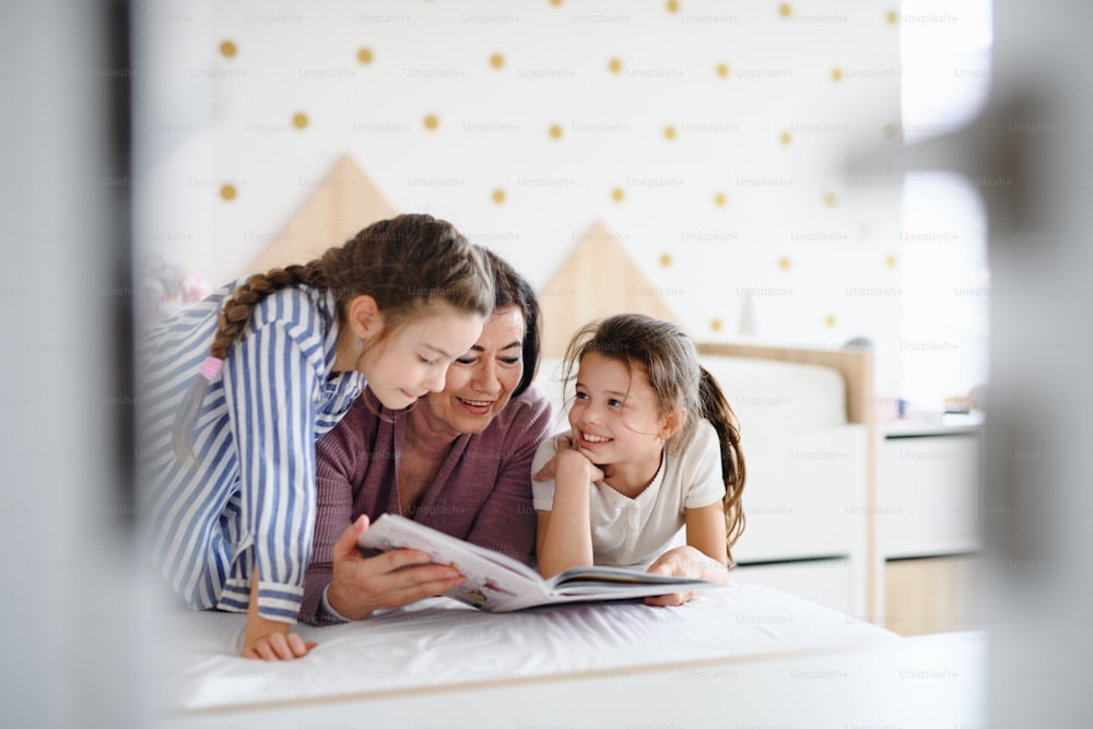 Happy small girls with a senior grandmother indoors in bedroom at home, reading on bed