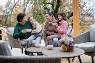 Two happy sisters with a mother and grandmother sitting and drinking tea outdoors in patio in autumn.