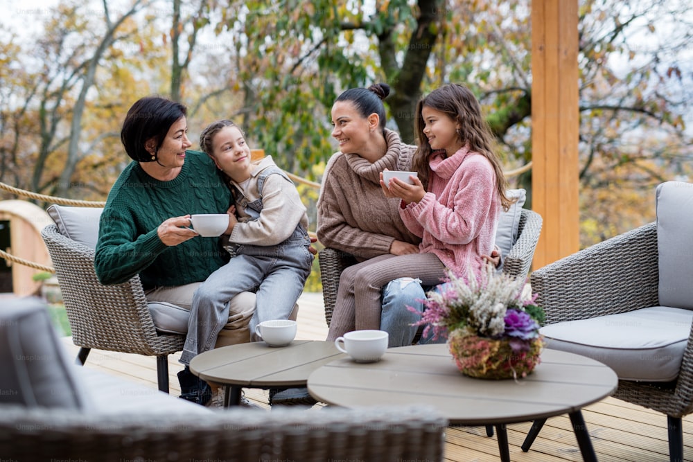 Two happy sisters with a mother and grandmother sitting and drinking tea outdoors in patio in autumn.
