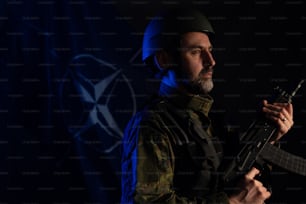 A soldier in military uniform and helmet holding weapon with NATO flag in background.