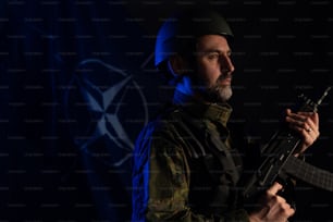 Ein Soldat in Militäruniform und Helm mit Waffe und NATO-Flagge im Hintergrund.