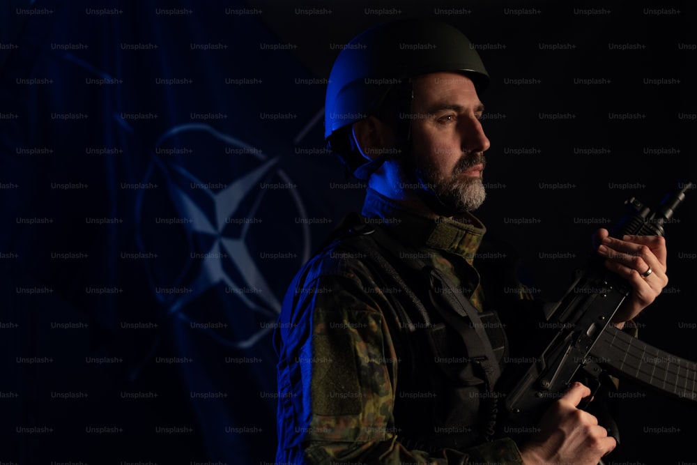 A soldier in military uniform and helmet holding weapon with NATO flag in background.