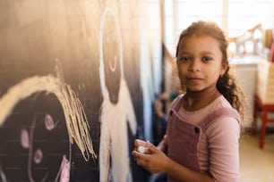 A little girl drawing with chalks on blackboard wall indoors in playroom, looking at camera.