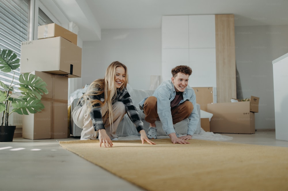 Una alegre pareja joven en su nuevo apartamento, extendiendo la alfombra. Concepción de la mudanza.