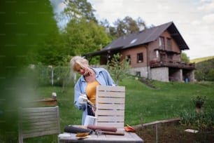 Una mujer mayor con un pincel impregnando una caja de madera al aire libre en el jardín.