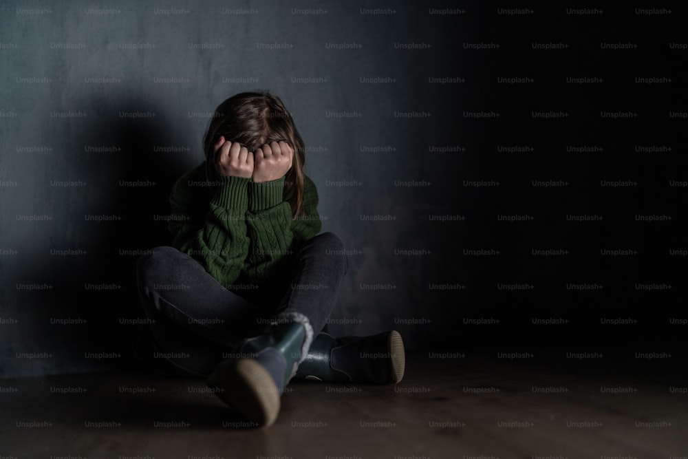 Una niña triste, sola en la oscuridad, sentada y con la cara cubierta.