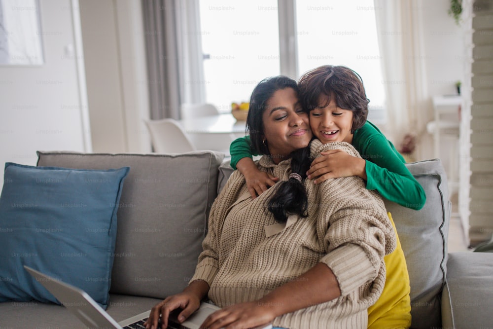 An Indian mother using laptop and her little son hugging herg on sofa at home.