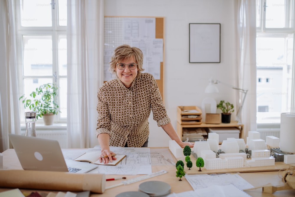 A mature woman architect with model of houses standing in office and looking at camera.