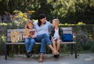 A happy senior woman with grandchildren sitting on bench and helping with homework outdoors in park.