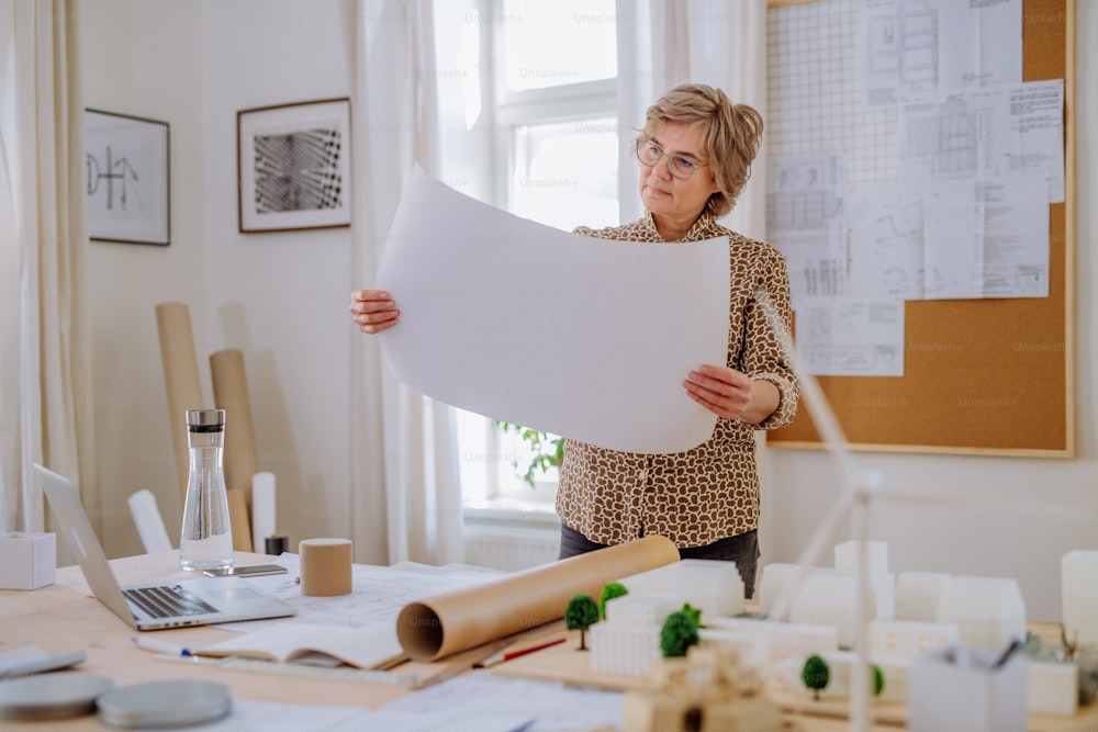A senior woman architect with model of houses looking at blueprints in office.