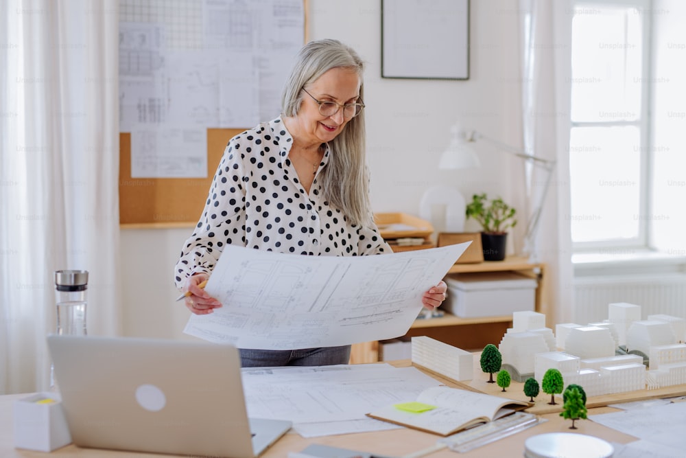 A senior woman architect with model of houses looking at blueprints in office.