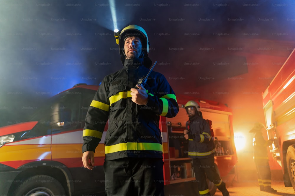 Une vue en contre-plongée d’un pompier parlant à un talkie-walkie avec un camion de pompiers en arrière-plan la nuit.