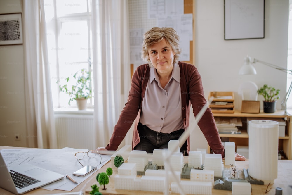 Happy senior woman architect with model of houses standing in office and looking at camera.