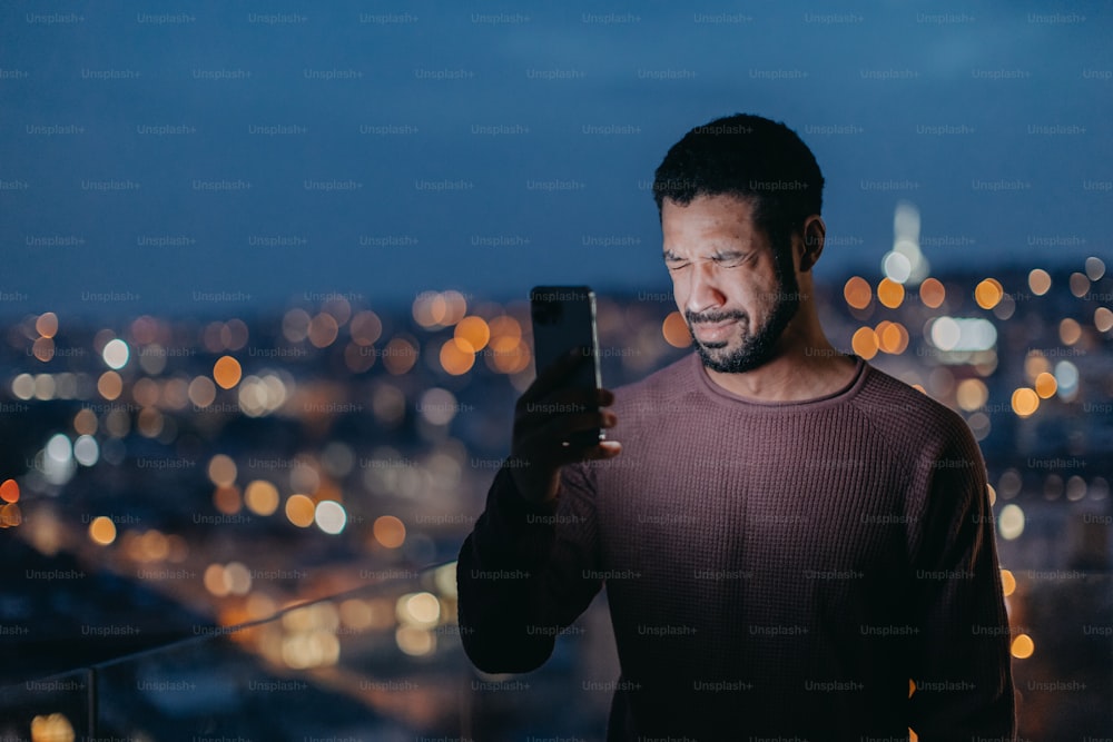 Un jeune homme avec un téléphone intelligent pleurant et debout sur le balcon avec vue sur la ville le soir à la maison.