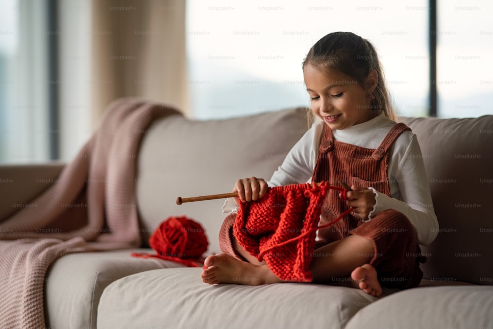Ein kleines Mädchen, das auf dem Sofa sitzt und zu Hause drinnen stricken lernt.