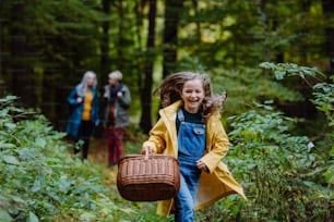 A happy little girl with basket running during walk with mother and grandmother outdoors in forest