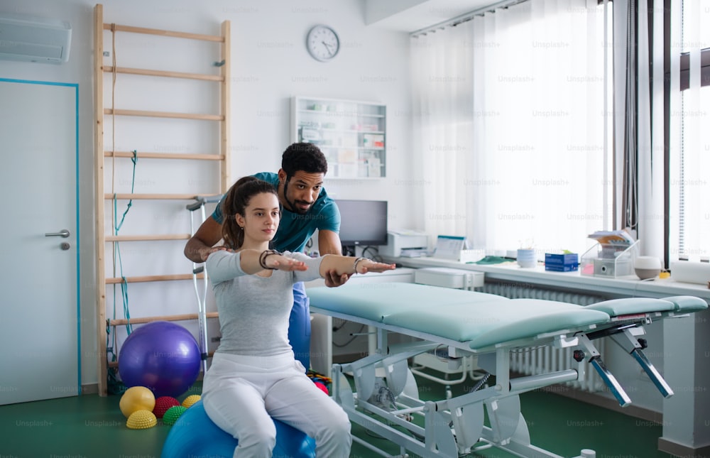 젊은 남성 물리 치료사가 물리학실에서 공을 타고 젊은 여성 환자와 함께 운동하고 있다