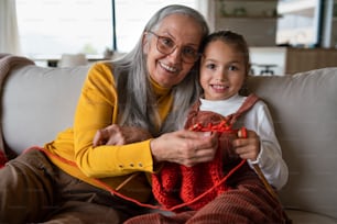 Une petite fille assise sur un canapé avec sa grand-mère et apprenant à tricoter à l’intérieur à la maison.