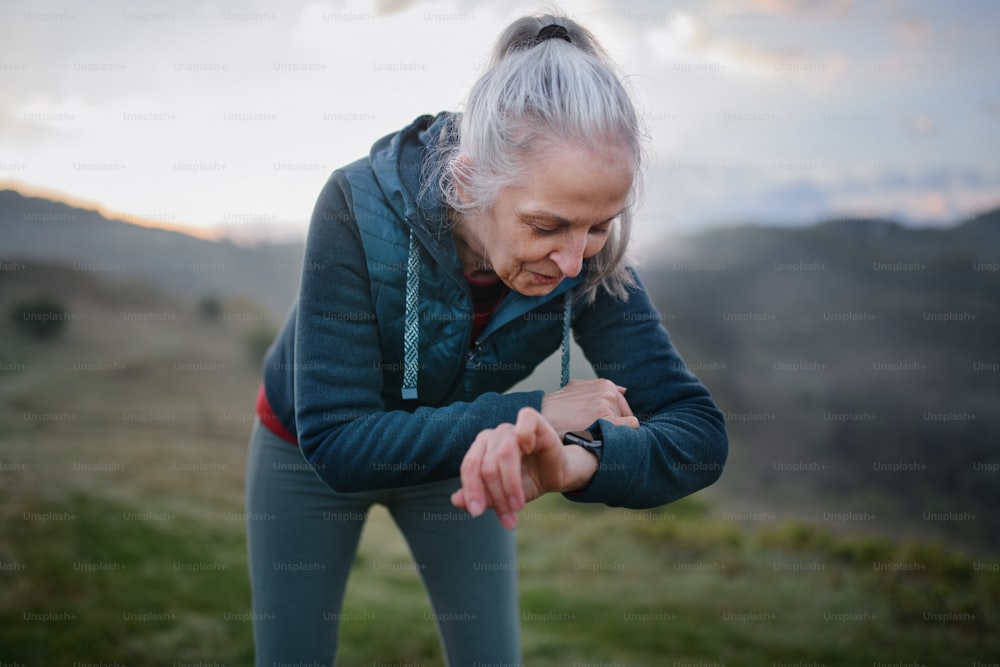 Una mujer mayor que corre y mira un reloj inteligente deportivo, verifica su rendimiento en la naturaleza temprano en la mañana con niebla y montañas en el fondo.
