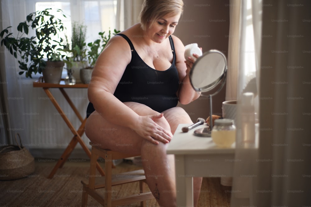 Una donna grassa seduta e che applica la crema sulle gambe a casa, concetto di cura di sé.