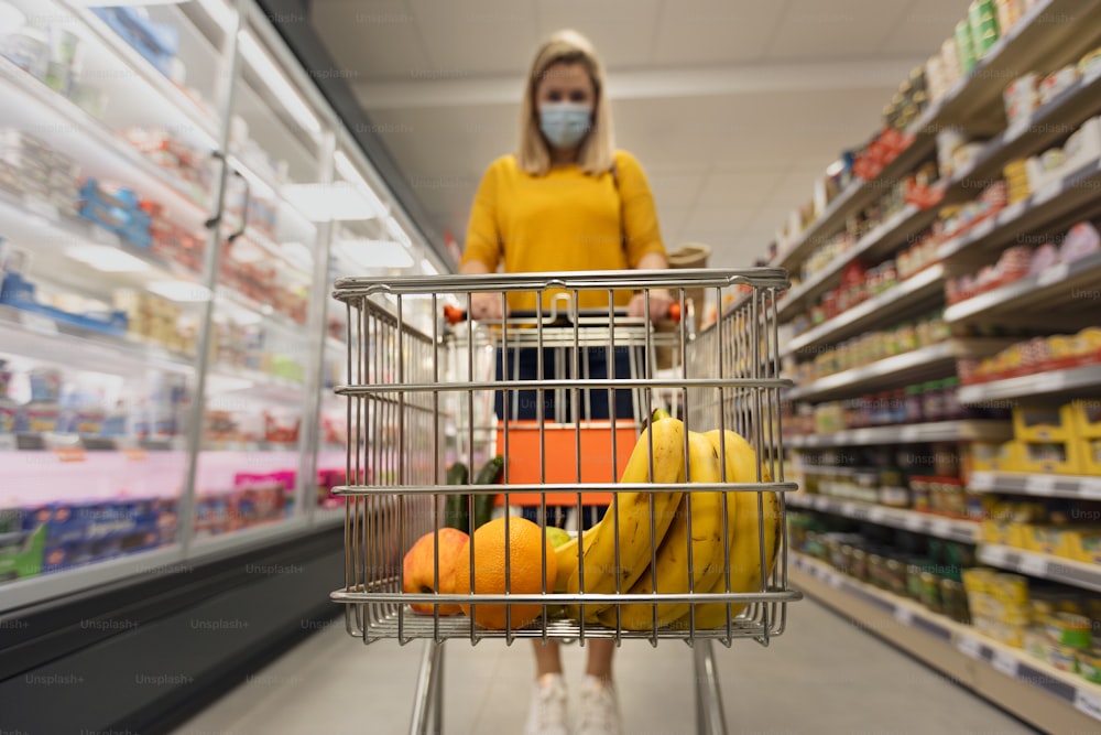 Una giovane donna in maschera facciale con carrello della spesa al supermercato, concetto di inflazione.