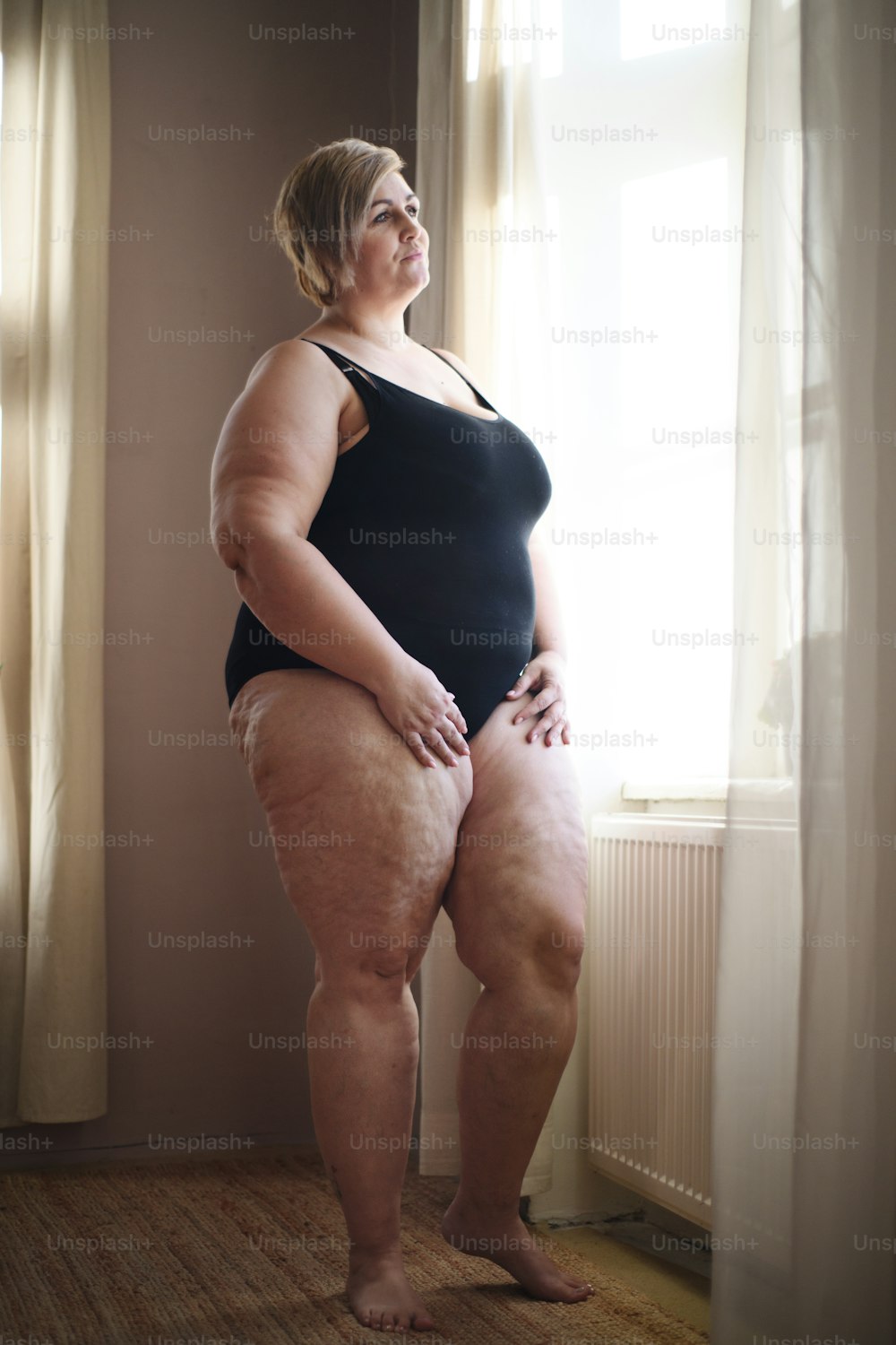 Depressed lonely overweight woman standing and looking through the window at home.