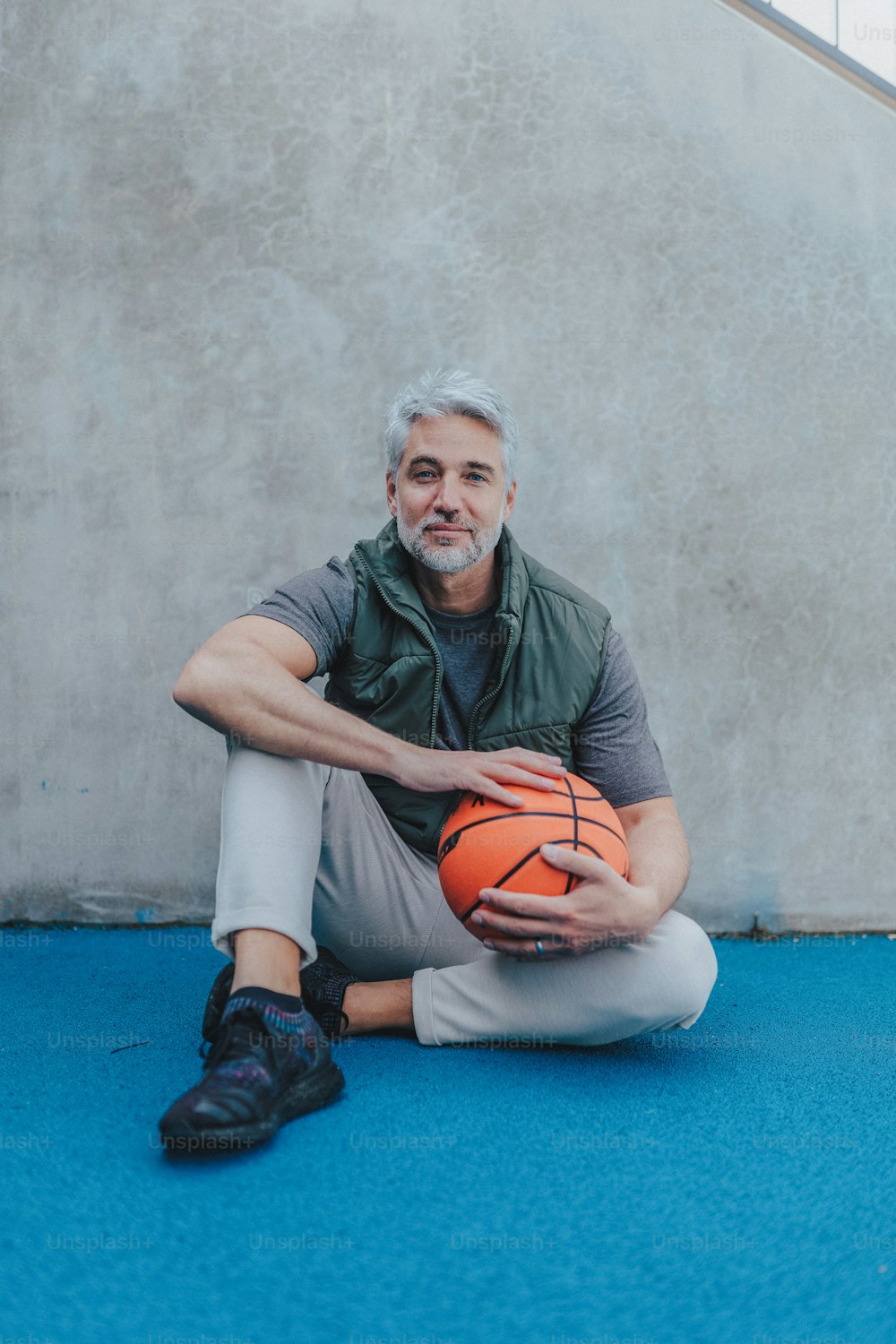 A mature man with baskatball sitting on courtoutdoors and looking at camera.