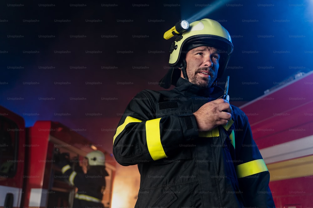 A low angle view of m firefighter talking to walkie talkie with fire truck in background at night.