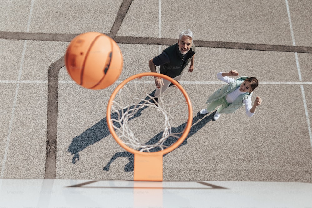 Um pai feliz e uma filha adolescente abraçados e de pé sob uma rede de aro de basquete, diretamente acima