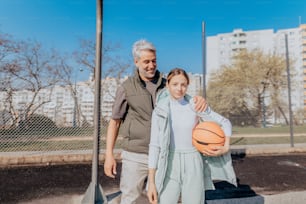 Ein glücklicher Vater und eine jugendliche Tochter, die sich umarmen und draußen auf dem Basketballplatz in die Kamera schauen.