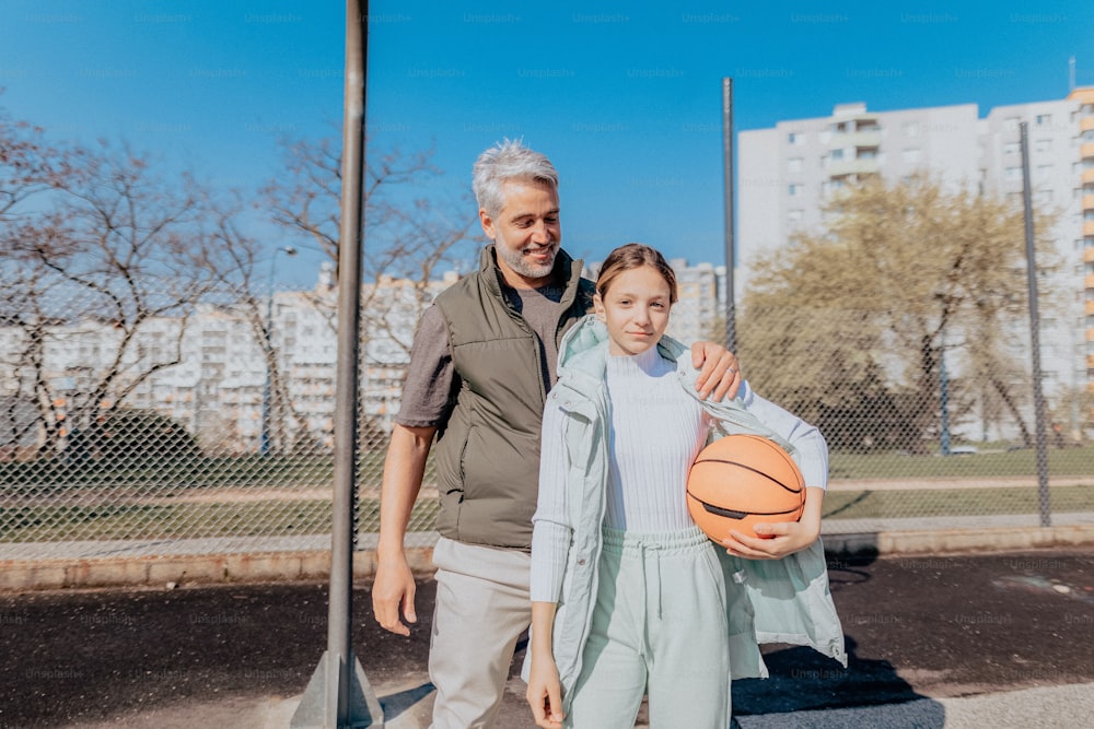Un père heureux et sa fille adolescente s’embrassent et regardent la caméra à l’extérieur sur le terrain de basket-ball.