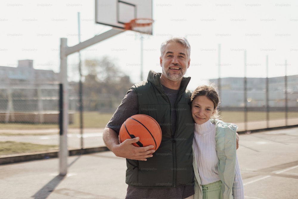 Um pai feliz e uma filha adolescente abraçados e olhando para a câmera do lado de fora da quadra de basquete.