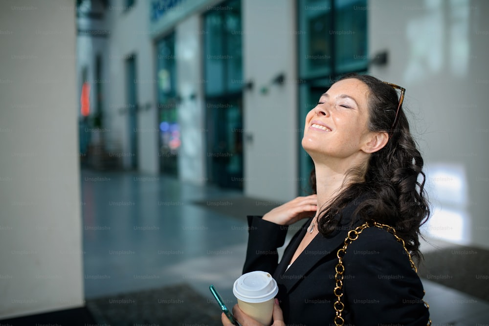 Eine erfolgreiche, glückliche Geschäftsfrau, die morgens in der Stadtstraße pendelt, Kaffeebecher hält und sich sonnt.