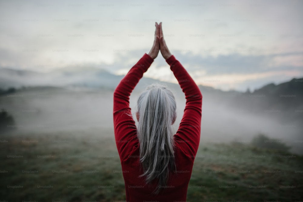A rear view of senior woman doing breathing exercise in nature on early morning with fog and mountains in background.