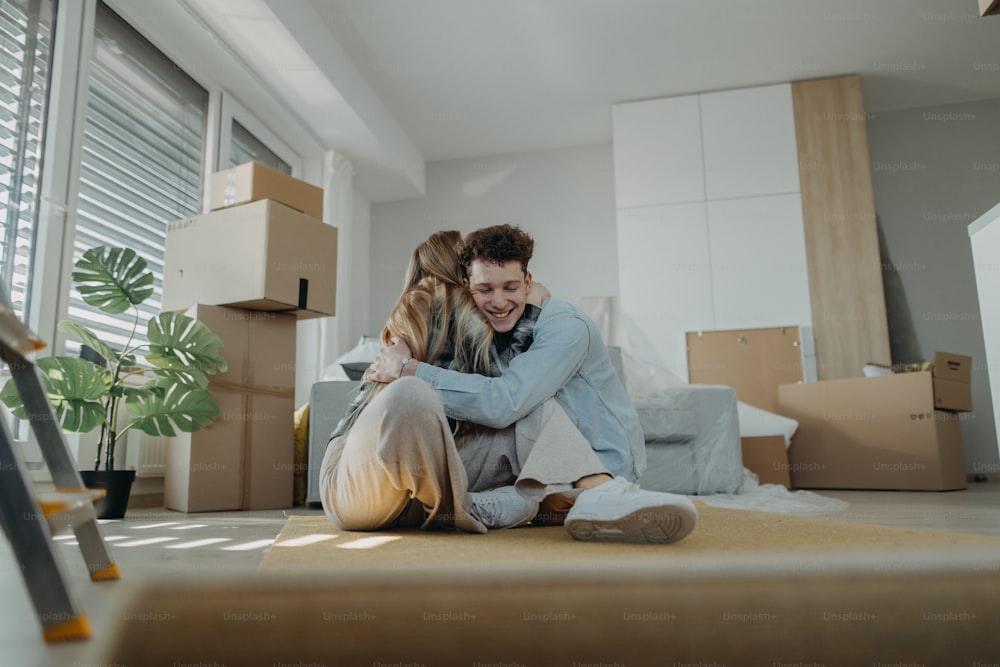 A cheerful young couple in their new apartment, sitting on floor and hugging. Conception of moving.