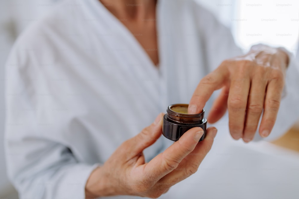 Una bella donna anziana in accappatoio che applica una crema naturale per il viso in bagno, nella cura della pelle e nel concetto di routine mattutina.
