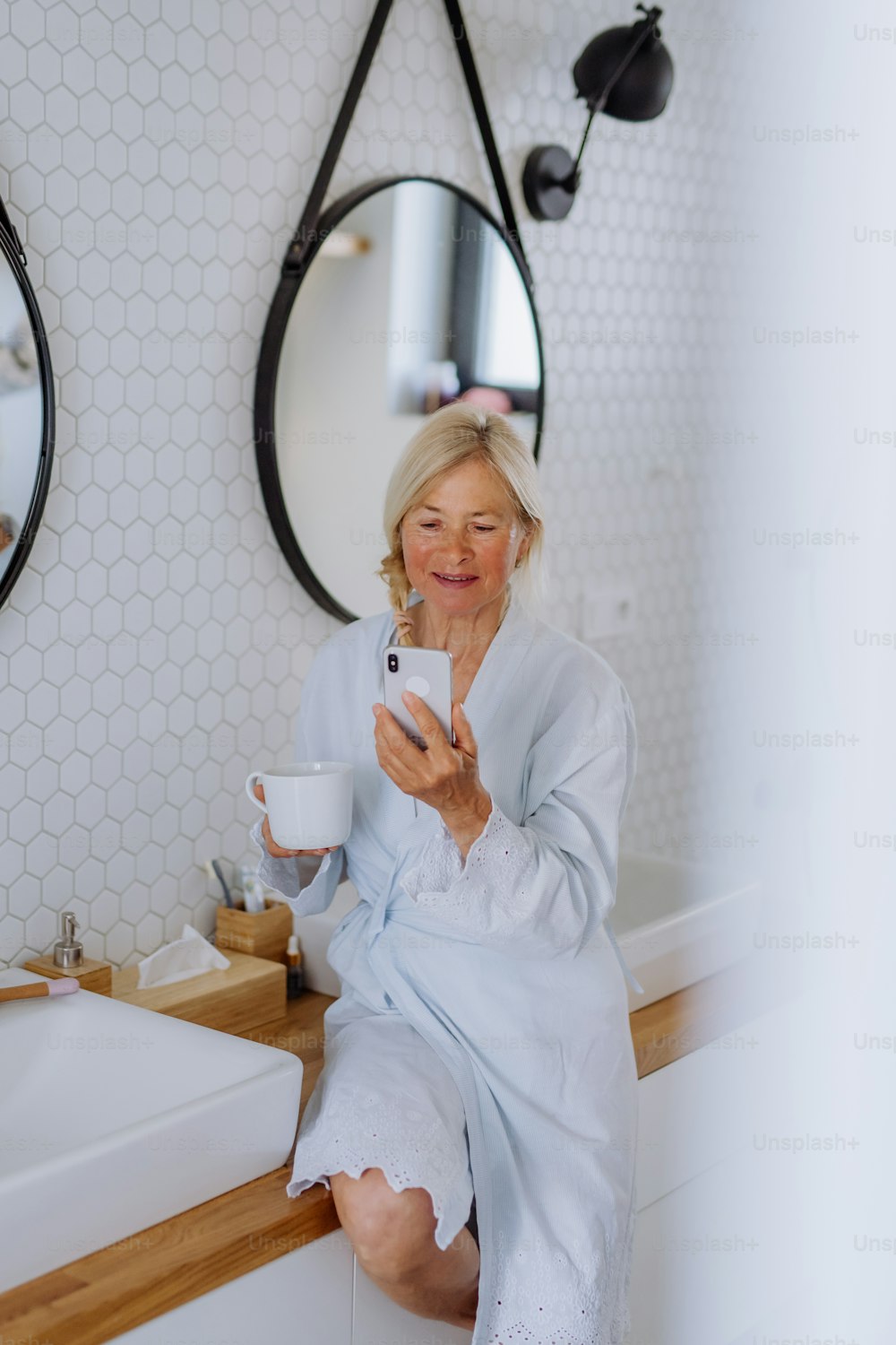 Una hermosa mujer mayor en bata de baño bebiendo té y usando un teléfono inteligente en el baño, concepto de relajación y bienestar.