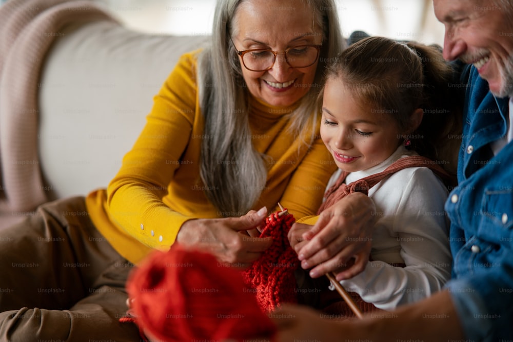 Une petite fille assise sur un canapé avec ses grands-parents et apprenant à tricoter à l’intérieur à la maison.