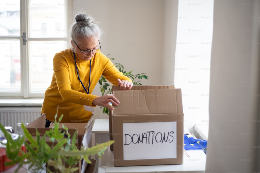 A senior volunteer woman packing box with Humanitarian aid for Ukrainian refugees in office