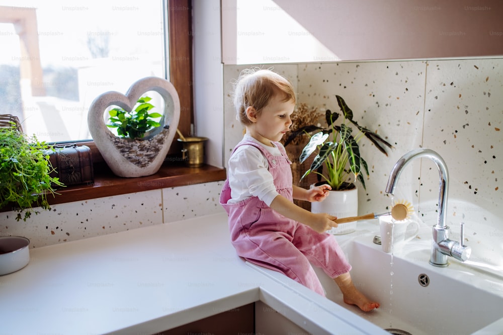 A little girl sitting on kitchen counter and washing cup in sink in kitchen.