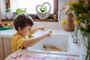 A little boy washing cup in sink in kitchen with wooden scrub, sustainable lifestlye.