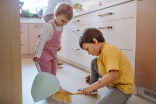 Um menino e uma menina ajudando a limpar a casa usando panela e escova enquanto varrem a sujeira do chão.