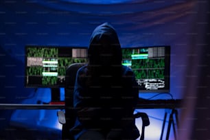 A hooded anonymous hacker by computer in the dark room at night, cyberwar concept.