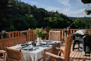 A dining table with wooden chairs set for dinner on the terrace with grill in summer, garden party. concept.