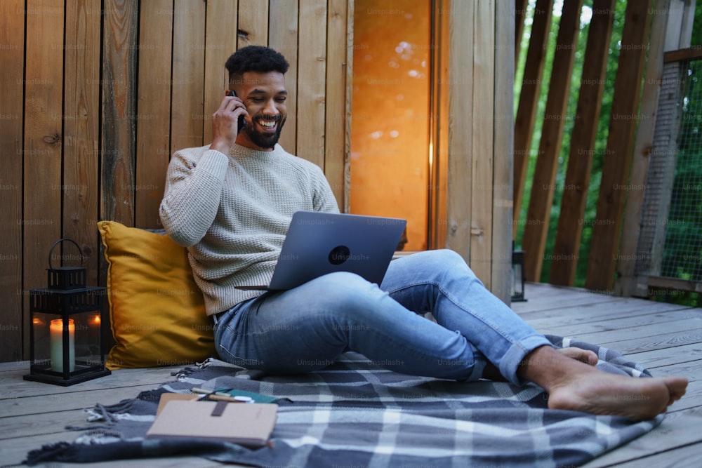 Un jeune homme heureux avec un ordinateur portable se reposant à l’extérieur dans une cabane dans les arbres, un week-end et un concept de bureau à distance.
