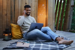 노트북을 들고 나무 위의 집에서 야외에서 쉬고 있는 행복한 청년, 주말 외출 및 원격 사무실 개념.