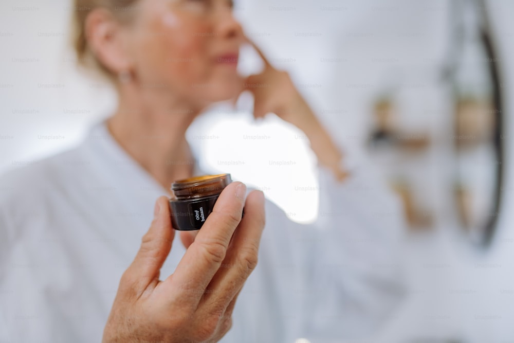 A beautiful senior woman in bathrobe applying natural face cream in bathroom, skin care and morning routine concept.