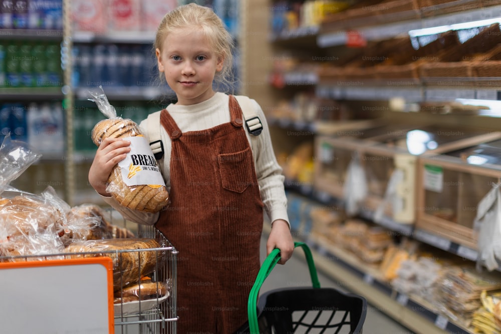Little girl standing in the middle of supermarket and buying bread, holding shopping basket.