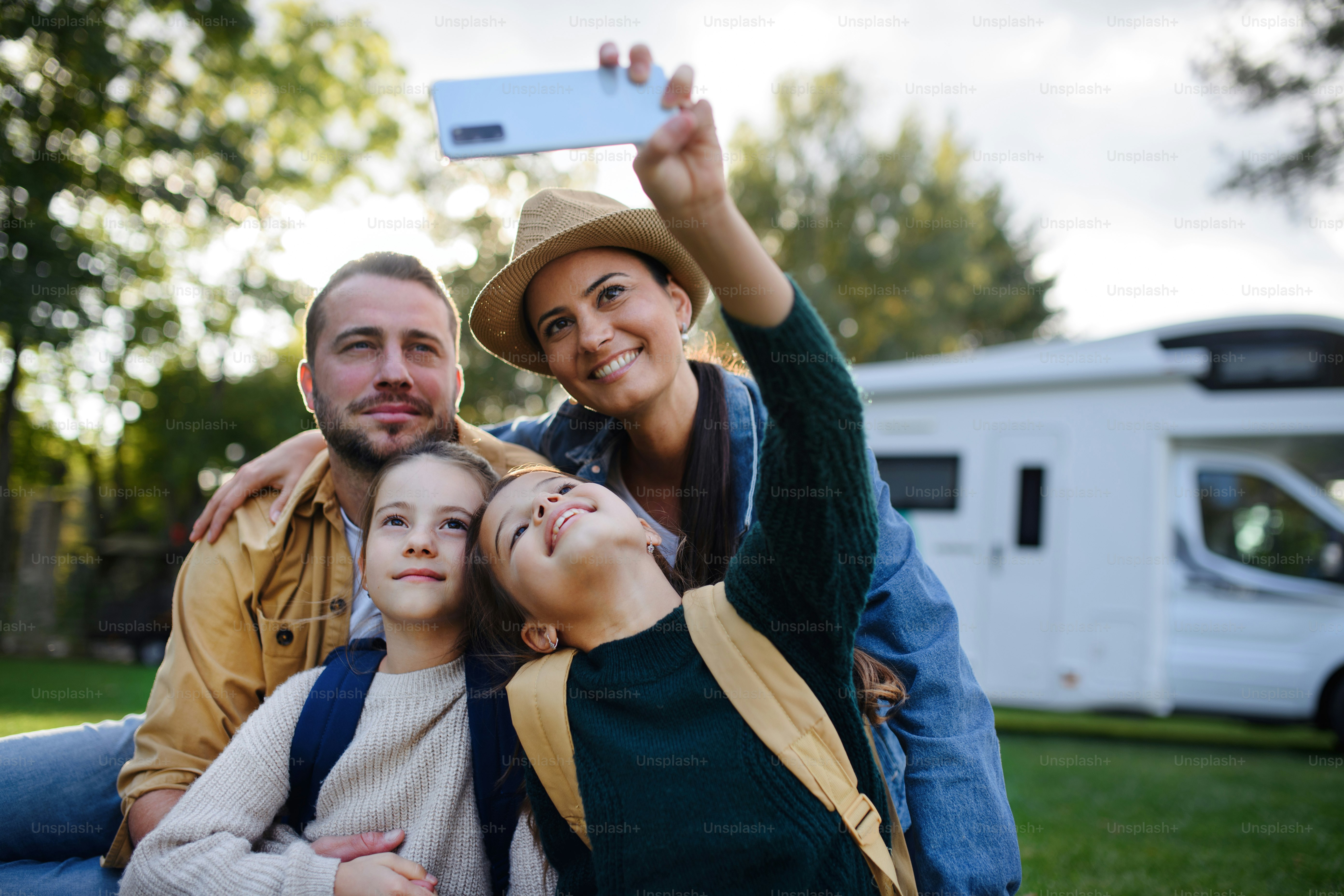 Une jeune famille heureuse avec deux enfants prenant un selfie avec une caravane à l’arrière-plan à l’extérieur.